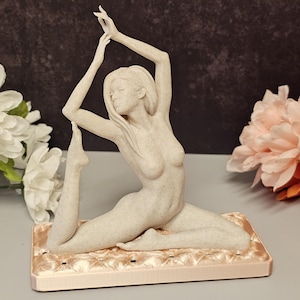 May include: A beige-colored yoga pose figurine. The sculpture depicts a woman in a seated yoga pose, with arms extended overhead and one leg raised. The figurine rests on a rectangular, peach-colored base with a quilted texture.