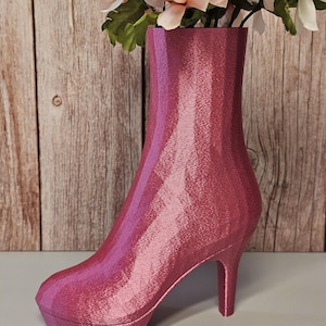 May include: A pink, high-heeled boot-shaped vase with a platform sole and stiletto heel. The vase is filled with artificial flowers in shades of pink, white, and green. The background is a wooden wall.