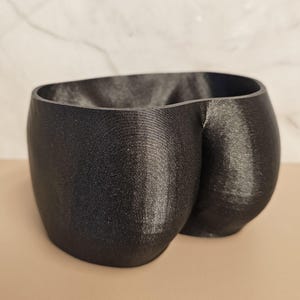 Op de afbeelding: Een zwarte, 3D-geprinte plantenbak in de vorm van een paar billen. De plantenbak heeft een getextureerd oppervlak en een brede opening. Het ontwerp is modern en uniek, geschikt voor kleine planten of vetplanten. De plantenbak is gemaakt van een donker, mat materiaal.