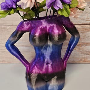 Op de afbeelding: Een decoratieve vaas in de vorm van een vrouwelijk torso, beschilderd in blauwe, paarse en zwarte tinten, met kunstbloemen. De bloemen zijn paars en perzik met groene bladeren. Unieke woondecoratie.