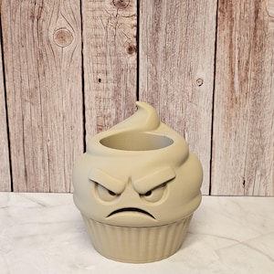 May include: A light beige cupcake-shaped container with an angry face design. The container has a detailed cupcake base and a swirl-shaped top, with a recessed area for holding items. The face features furrowed eyebrows and a downturned mouth.