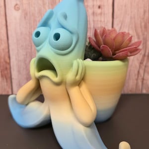 May include: A 3D printed planter shaped like a shocked banana with a gradient color scheme transitioning from blue at the top to yellow at the base. The banana figure has wide eyes, a gaping mouth, and hands pressed against its cheeks, holding a succulent.