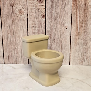 May include: A beige ceramic toilet figurine with a lid, bowl, and base. The miniature toilet is a decorative item, likely for a dollhouse or novelty collection. The background is a wooden wall.