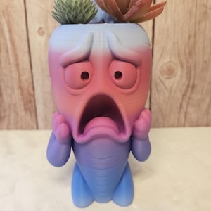 May include: A 3D printed planter shaped like a character with a wide-eyed, shocked expression, featuring a gradient colour transition from light blue at the base to pink and white at the top, with two small hands touching its face and a succulent.