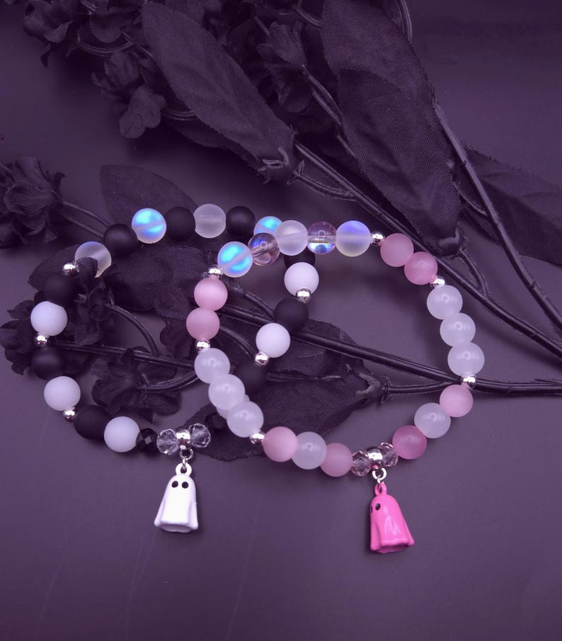 Op de afbeelding: Drie armbanden met kralen met een Halloween-thema. De armbanden hebben zwarte, witte, roze en iriserende kralen. Elke armband heeft een spookbedel. De achtergrond is donkerpaars.