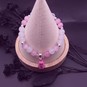 Op de afbeelding: Een armband met afwisselend roze en witte kralen, geaccentueerd met kleine zilverkleurige elementen. Een roze spookbedel hangt in het midden. De armband wordt gepresenteerd op een houten standaard met een lichtgekleurde stof.