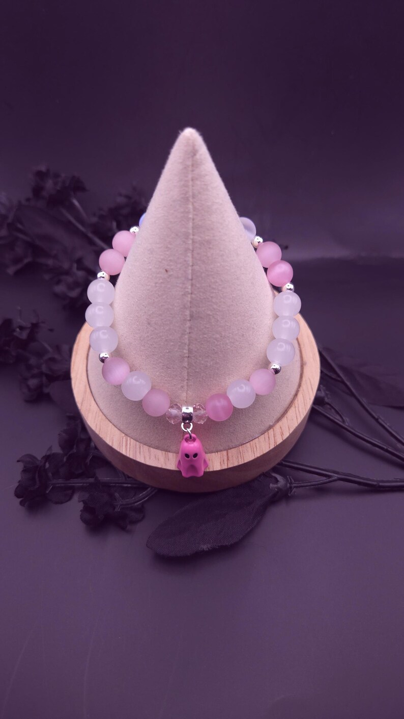 Op de afbeelding: Een armband met afwisselend roze en witte kralen, geaccentueerd met kleine zilverkleurige elementen. Een roze spookbedel hangt in het midden. De armband wordt gepresenteerd op een houten standaard met een lichtgekleurde stof.