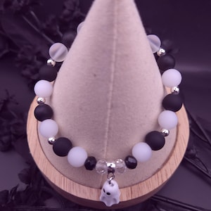 Op de afbeelding: Een Halloween-armband met afwisselend matzwarte en witte kralen, geaccentueerd met zilverkleurige afstandhouders. Een kleine, witte spookbedel hangt in het midden. De armband wordt gepresenteerd op een lichtgekleurde, kegelvormige standaard.