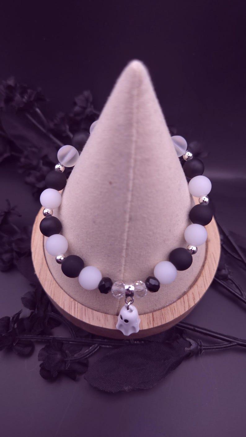 Op de afbeelding: Een Halloween-armband met afwisselend matzwarte en witte kralen, geaccentueerd met zilverkleurige afstandhouders. Een kleine, witte spookbedel hangt in het midden. De armband wordt gepresenteerd op een lichtgekleurde, kegelvormige standaard.