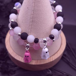 Op de afbeelding: Twee armbanden met een Halloween-thema. De ene armband heeft zwarte, witte en roze kralen. De andere armband heeft roze, witte en transparante kralen. Elke armband heeft een spookbedel. De armbanden worden op een houten standaard getoond.
