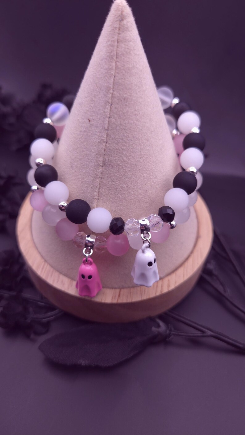 Op de afbeelding: Twee armbanden met een Halloween-thema. De ene armband heeft zwarte, witte en roze kralen. De andere armband heeft roze, witte en transparante kralen. Elke armband heeft een spookbedel. De armbanden worden op een houten standaard getoond.