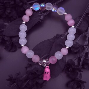 Op de afbeelding: Een armband met roze en iriserende kralen, versierd met een kleine roze spookbedel. De kralen zijn geregen op een elastische band. De armband staat op een donkerpaarse achtergrond met zwarte bloemaccenten.