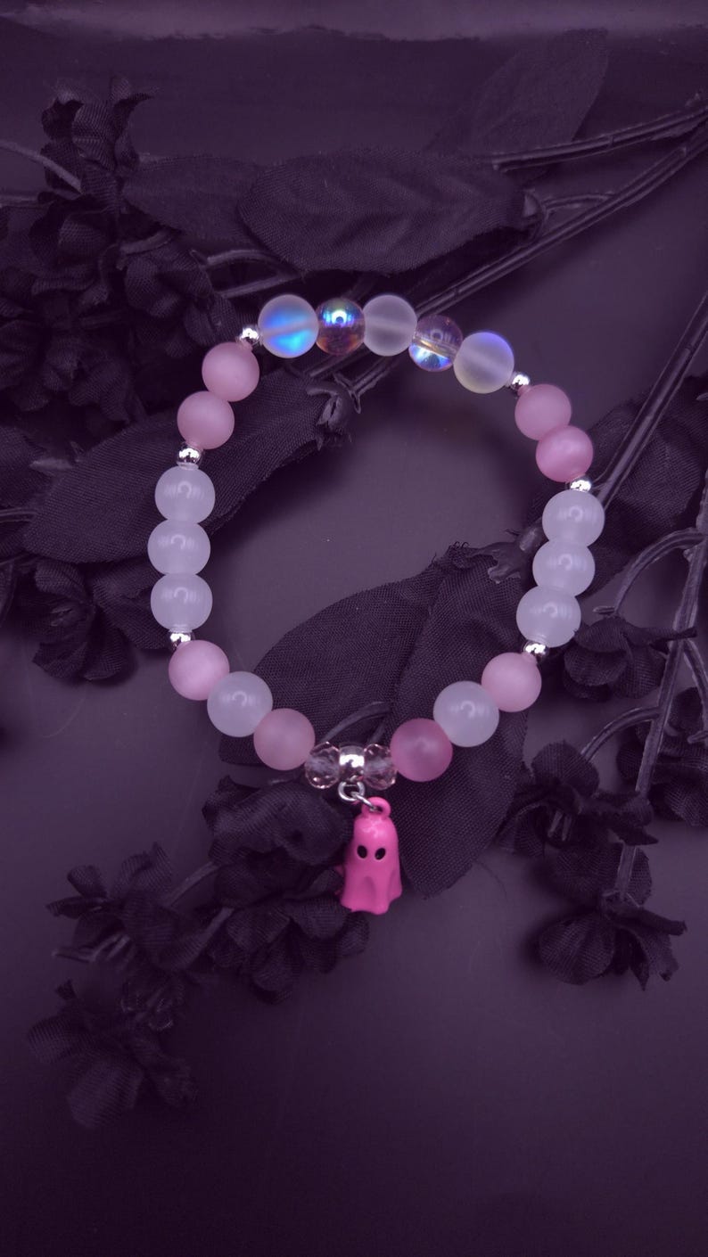 Op de afbeelding: Een armband met roze en iriserende kralen, versierd met een kleine roze spookbedel. De kralen zijn geregen op een elastische band. De armband staat op een donkerpaarse achtergrond met zwarte bloemaccenten.