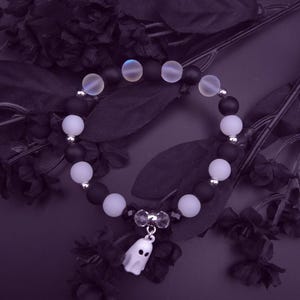 Op de afbeelding: Een armband met zwarte en witte kralen, afgewisseld met iriserende kralen. Een kleine witte spookbedel hangt in het midden. De armband is geplaatst tegen een donkerpaarse achtergrond met zwarte bloemaccenten.
