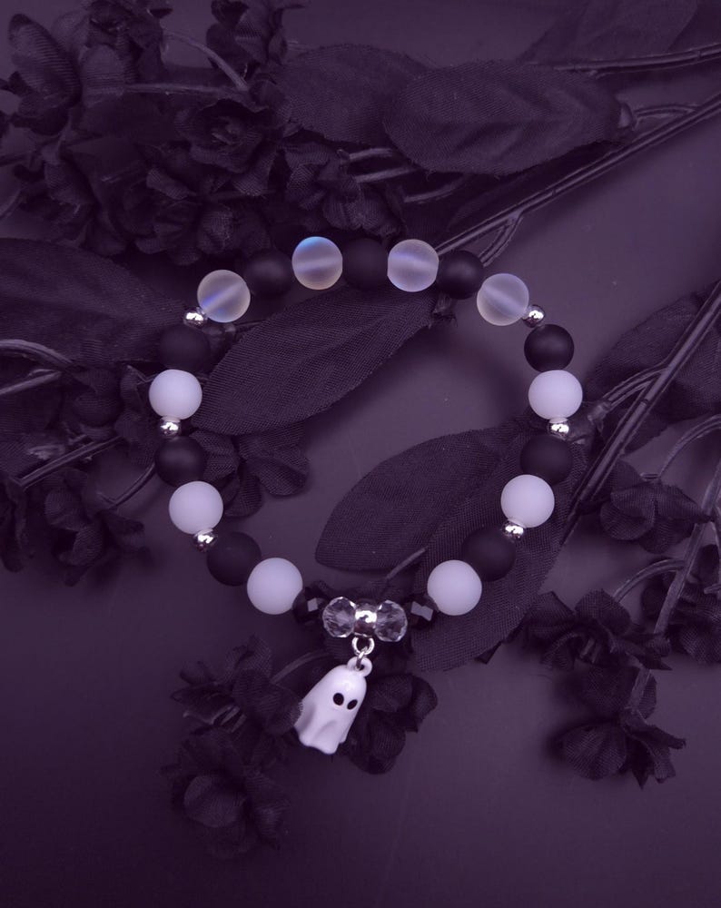 Op de afbeelding: Een armband met zwarte en witte kralen, afgewisseld met iriserende kralen. Een kleine witte spookbedel hangt in het midden. De armband is geplaatst tegen een donkerpaarse achtergrond met zwarte bloemaccenten.