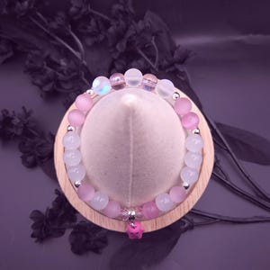 Op de afbeelding: Een armband met roze en witte kralen, met zilverkleurige accenten. Een kleine roze bedel hangt in het midden. De armband wordt gepresenteerd op een houten standaard tegen een donkerpaarse achtergrond.