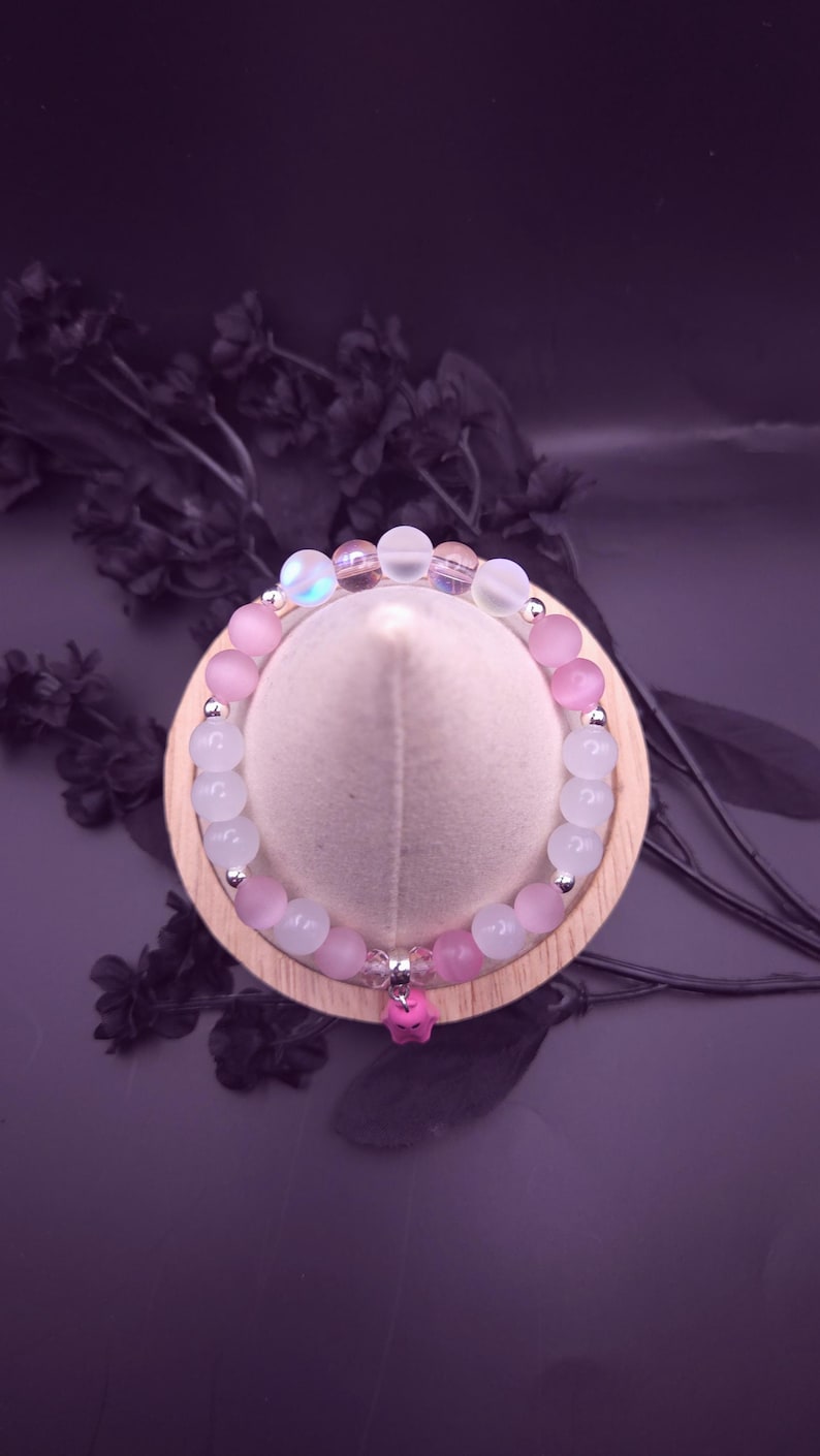 Op de afbeelding: Een armband met roze en witte kralen, met zilverkleurige accenten. Een kleine roze bedel hangt in het midden. De armband wordt gepresenteerd op een houten standaard tegen een donkerpaarse achtergrond.