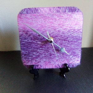 Puede incluir: Un reloj cuadrado con una esfera envuelta en hilo morado y rosa. El reloj tiene manecillas plateadas y está sostenido por un soporte negro. El hilo crea una apariencia texturizada y hecha a mano. El reloj es un artículo decorativo.