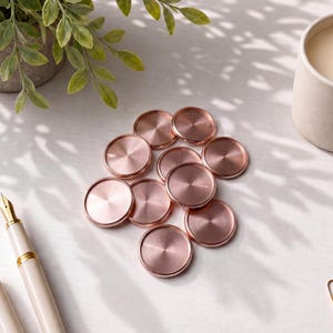 May include: A collection of eleven circular rose gold metal discs with a brushed finish, arranged in a cluster. The discs feature a smooth, flat surface with a slightly raised outer rim, reflecting light across their metallic, copper-toned circular faces.