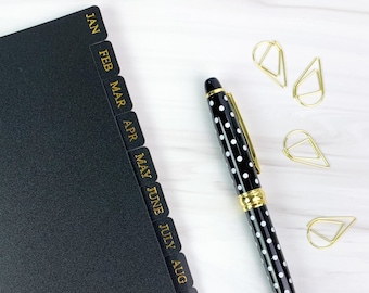 May include: A black planner with gold monthly tabs, a black and white polka dot pen, and gold paper clips on a white surface.