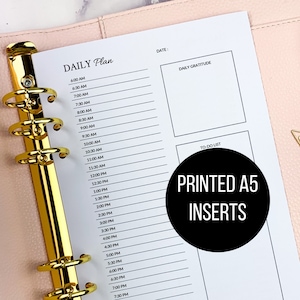 May include: A white A5 planner insert with a daily schedule from 6:00 AM to 9:00 PM, a section for daily gratitude, and a to-do list. The text "PRINTED A5 INSERTS" is printed in black on a white circle.