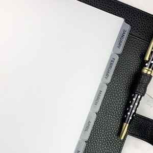 May include: A planner with monthly dividers labeled January through June. The dividers are gray with white text. A black and white polka dot pen with gold accents rests on the black leather planner.