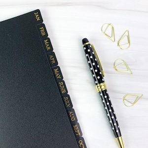 May include: A black planner with gold monthly tabs, a black and white polka dot pen, and gold paper clips on a white surface.