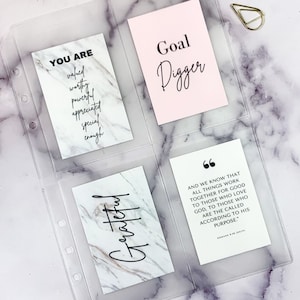 May include: Clear plastic binder with four inspirational cards. The cards have marble and pink backgrounds with black text that reads "YOU ARE", "Goal Digger", "Grateful", and a Bible verse. Gold paper clips are in the background.