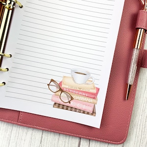 May include: A white planner with lined pages and a gold binder. The planner is open to a page with a watercolor illustration of a stack of books, a cup of coffee, and a pair of glasses. The books have the names of designer brands on them, including "Emporio Armani", "Burberry", "Dolce & Gabbana", "Gucci", and "Bvlgari".