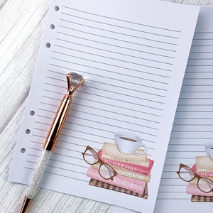 May include: A white lined notebook with a pink and brown illustration of a stack of books with a cup of coffee and glasses on top. The text on the books reads "EMPORIO ARMANI", "BURBERRY", "GABBANA", "CCI", and "BVLGARI".