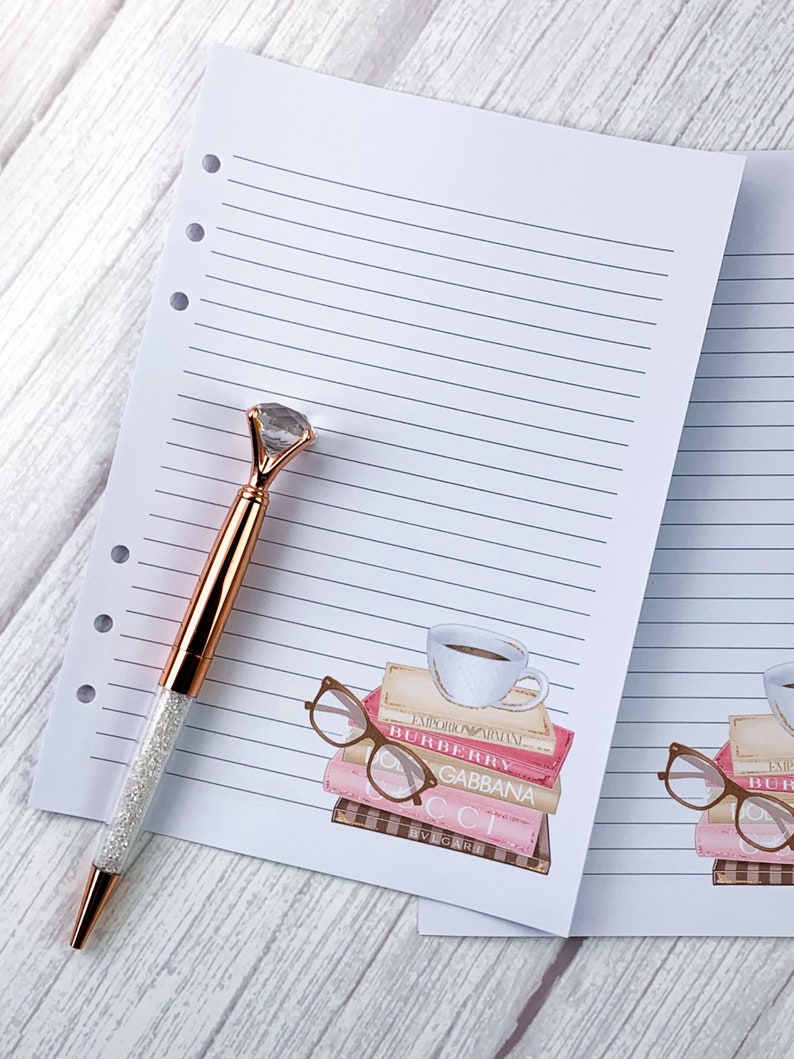 May include: A white lined notebook with a pink and brown illustration of a stack of books with a cup of coffee and glasses on top. The text on the books reads "EMPORIO ARMANI", "BURBERRY", "GABBANA", "CCI", and "BVLGARI".