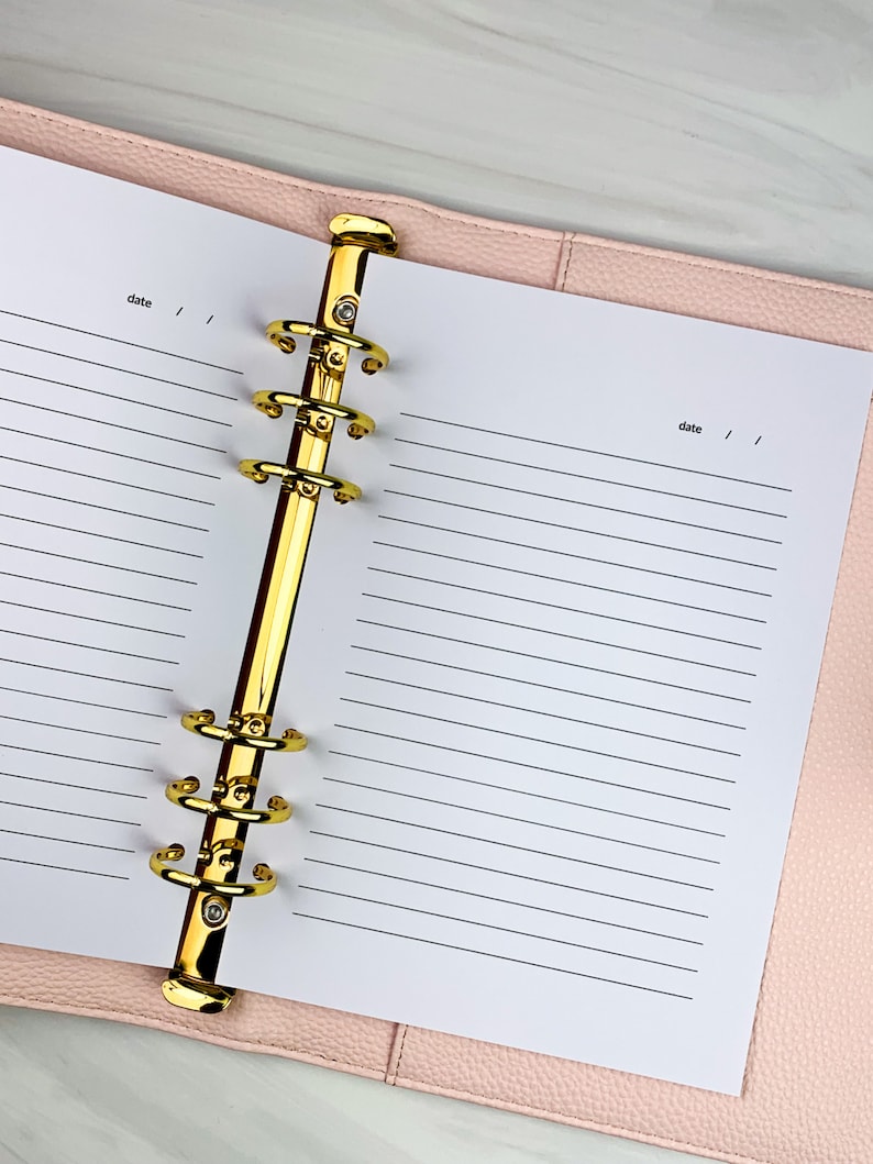 Pu&ograve; includere: Un'agenda rilegata in pelle rosa con anelli dorati e carta bianca rigata. L'agenda ha la parola "data" scritta su entrambi i lati delle pagine.