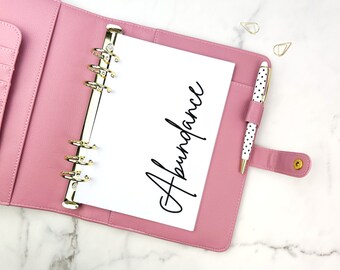 May include: A pink planner with gold accents, open to a page with the word "Abundance" in elegant script. A polka-dot pen rests in a holder on the right. Gold paperclips are scattered nearby. The planner is on a white marble surface.