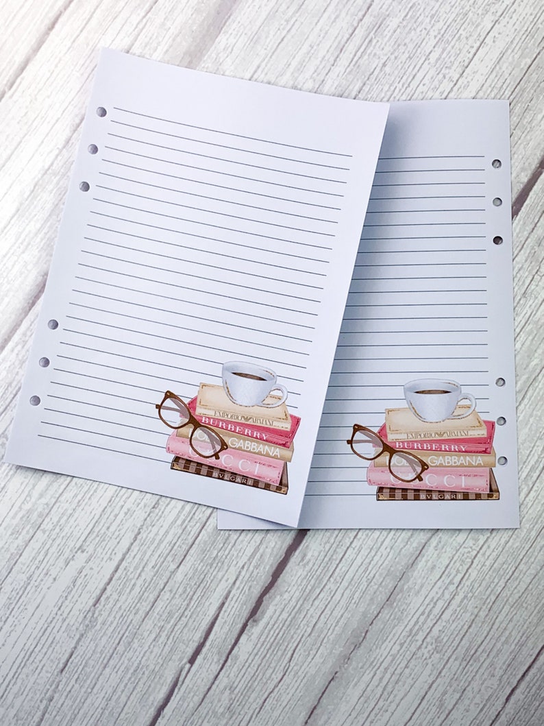 May include: Two white lined pages with holes on the left side. Each page has a stack of pink and brown books with a coffee cup on top and a pair of glasses next to the books. The books have the text "BURBERRY" and "GABBANA" on them.