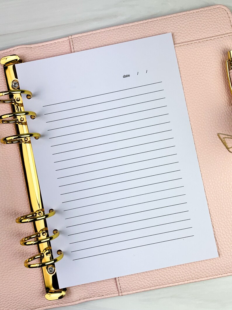Pu&ograve; includere: Un'agenda rilegata in pelle rosa con anelli dorati e una pagina bianca con righe aperta su una pagina bianca con la parola "data" scritta in alto.