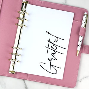 May include: A pink leather bound planner with gold rings and a white page that says "Grateful" in black script. A white pen with black polka dots is in the pen holder.
