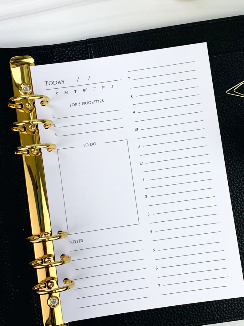 May include: A white daily planner page with a gold binder. The page has sections for "Today", "Top 3 Priorities", "To Do", and "Notes".