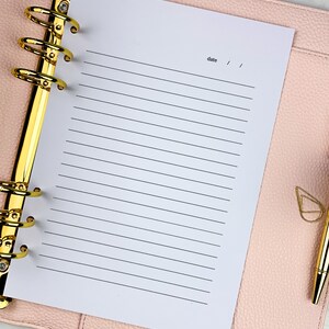 Pu&ograve; includere: Una pagina di carta bianca rigata in un planner rilegato in pelle rosa con anelli dorati. La pagina ha la parola "data" scritta in alto con due righe per la data.