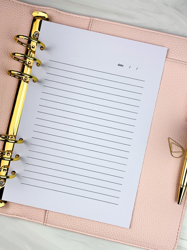 Pu&ograve; includere: Una pagina di carta bianca rigata in un planner rilegato in pelle rosa con anelli dorati. La pagina ha la parola "data" scritta in alto con due righe per la data.