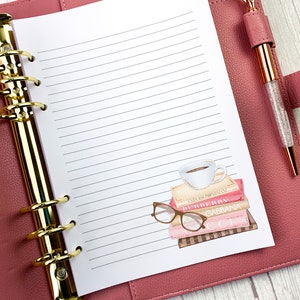 May include: A pink leather bound planner with gold rings and a white lined page. The page has a sticker of a stack of books with a cup of coffee on top and a pair of glasses on the side. The books have the names "Emporio Armani", "Burberry", "Dolce & Gabbana", "Gucci", and "Bvlgari" on the spines.