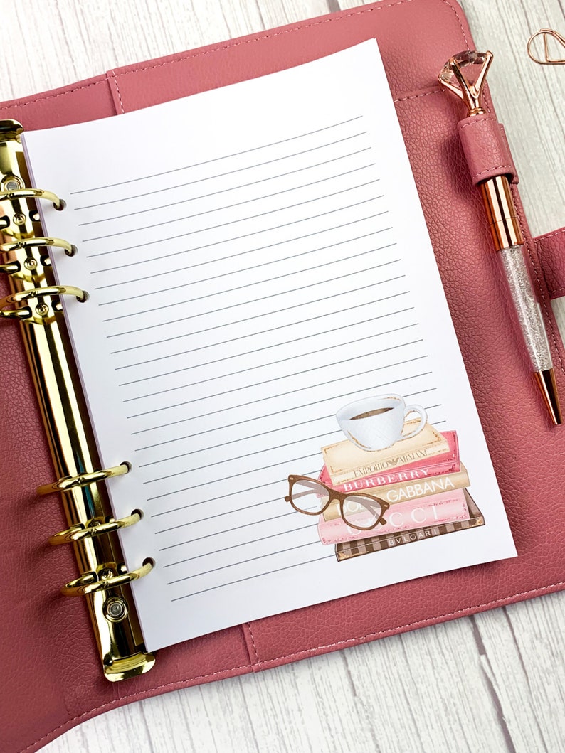 May include: A pink leather bound planner with gold rings and a white lined page. The page has a sticker of a stack of books with a cup of coffee on top and a pair of glasses on the side. The books have the names "Emporio Armani", "Burberry", "Dolce & Gabbana", "Gucci", and "Bvlgari" on the spines.