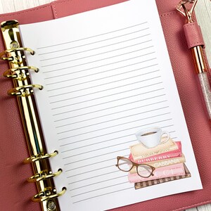 May include: A pink leather bound planner with gold rings and a white lined page. The page has a sticker of a stack of books with a coffee cup on top and a pair of glasses on the side. The books have the names of designer brands on them, including "Burberry", "Gucci", and "Dolce & Gabbana".