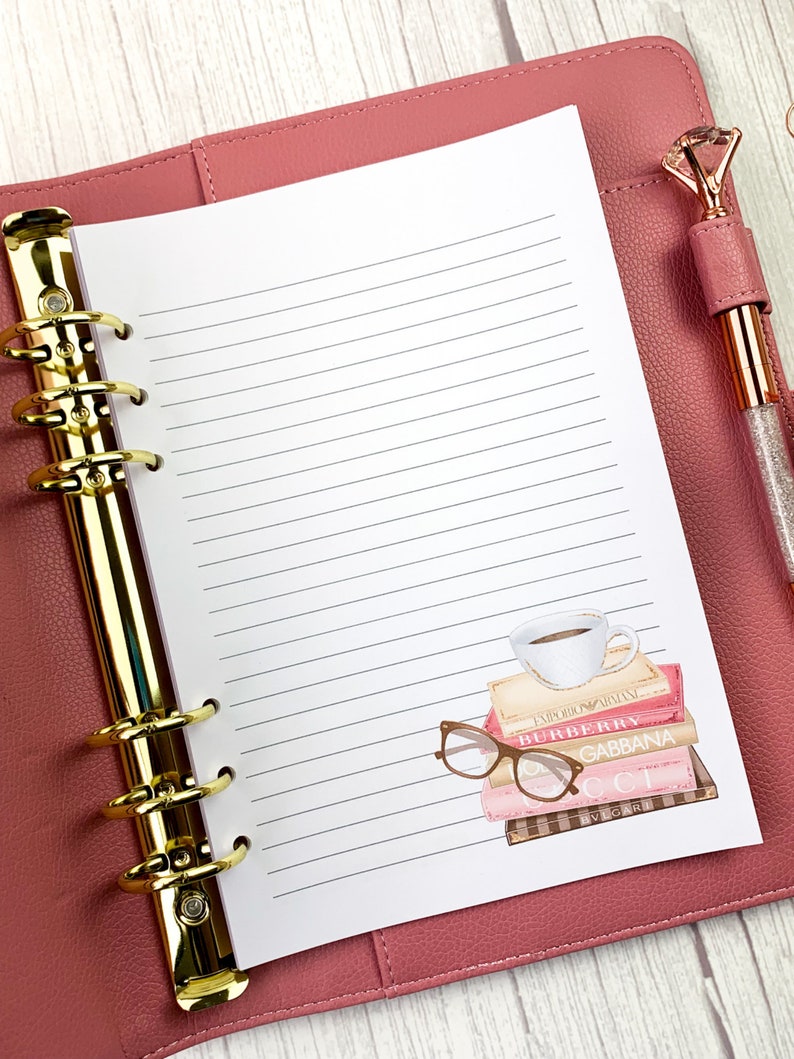 May include: A pink leather bound planner with gold rings and a white lined page. The page has a sticker of a stack of books with a coffee cup on top and a pair of glasses on the side. The books have the names of designer brands on them, including "Burberry", "Gucci", and "Dolce & Gabbana".