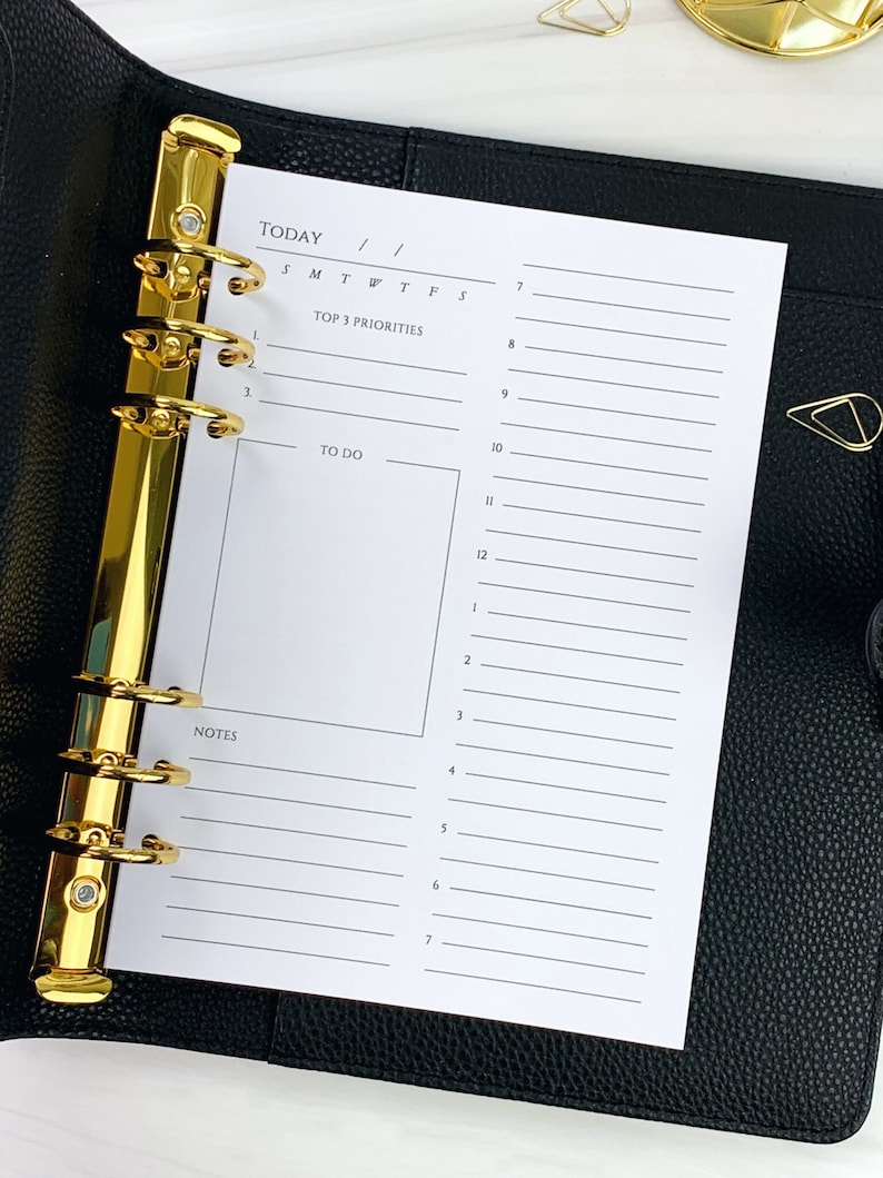 May include: A white daily planner page with a gold binder. The page has sections for "Today", "Top 3 Priorities", "To Do", and "Notes".
