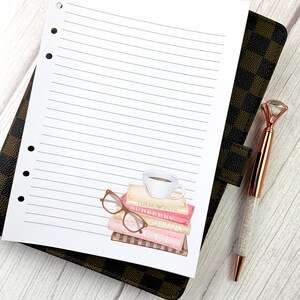 May include: A white lined notebook with a sticker of a stack of books with a coffee cup on top and a pair of glasses on the side. The books have the names of designer brands on the spines: "EMPORIO ARMANI", "BURBERRY", "GABBANA", "GUCCI", and "VERSACE".