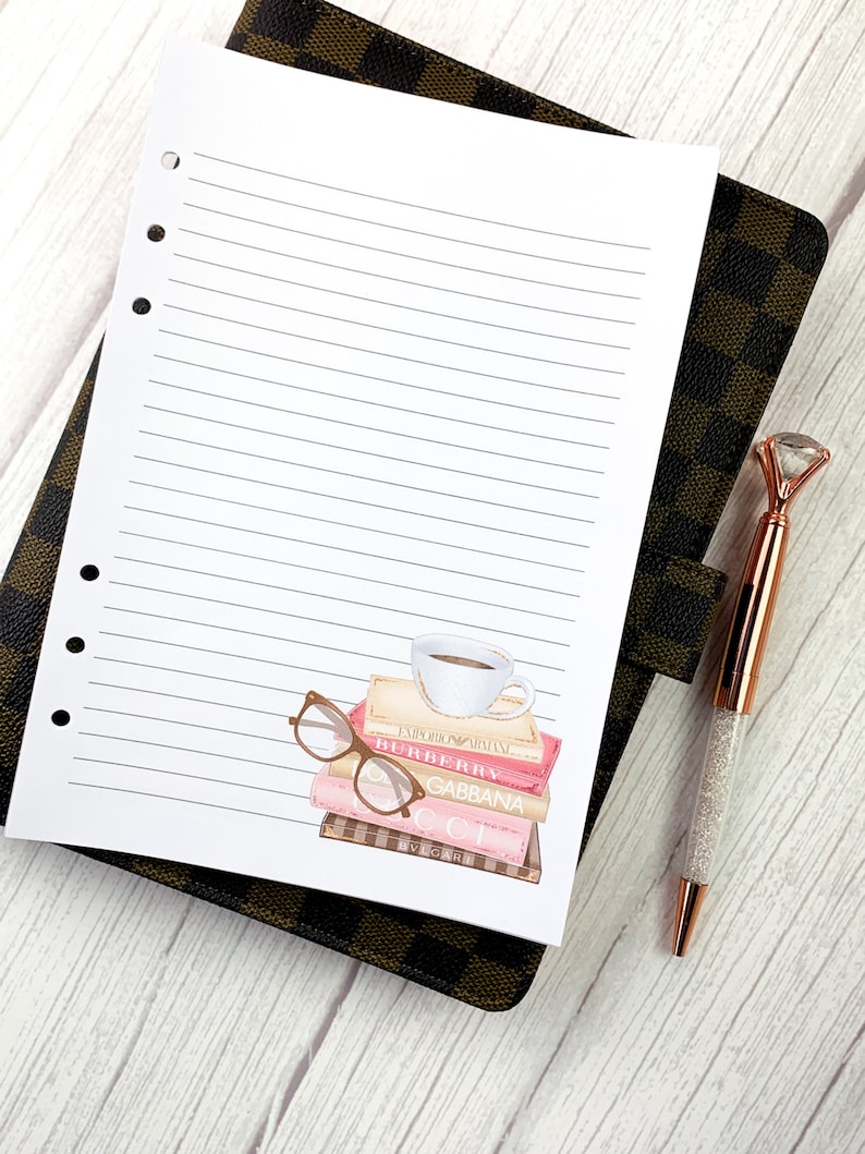 May include: A white lined notebook with a sticker of a stack of books with a coffee cup on top and a pair of glasses on the side. The books have the names of designer brands on the spines: "EMPORIO ARMANI", "BURBERRY", "GABBANA", "GUCCI", and "VERSACE".