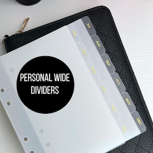May include: Clear plastic personal wide dividers with gold lettering for monthly organization. The dividers are labeled with the months of the year and are placed on top of a black quilted wallet. The text "PERSONAL WIDE DIVIDERS" is in a black circle.