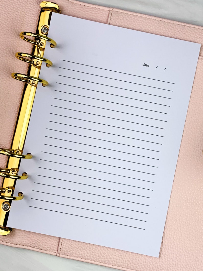 Pu&ograve; includere: Una pagina bianca con carta rigata in un'agenda rilegata in pelle rosa con anelli dorati. La pagina ha la parola "data" scritta in alto con uno spazio per la data.