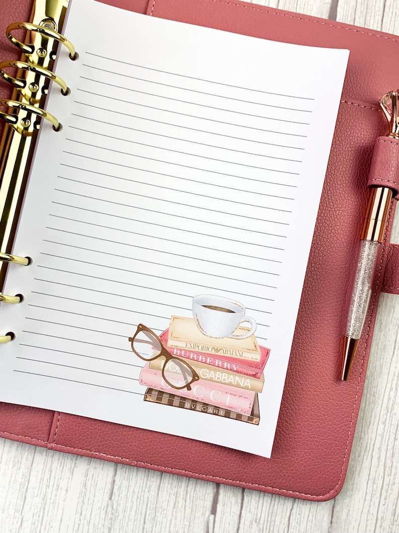 May include: A white planner with lined pages and a gold binder. The planner is open to a page with a watercolor illustration of a stack of books, a cup of coffee, and a pair of glasses. The books have the names of designer brands on them, including "Emporio Armani", "Burberry", "Dolce & Gabbana", "Gucci", and "Bvlgari".