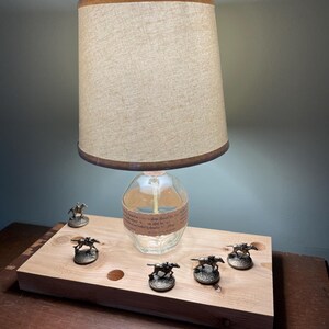 May include: A lamp made from a clear glass bottle with a beige lampshade. The bottle has a label with text. The lamp is on a wooden base with five small metal horse figurines.